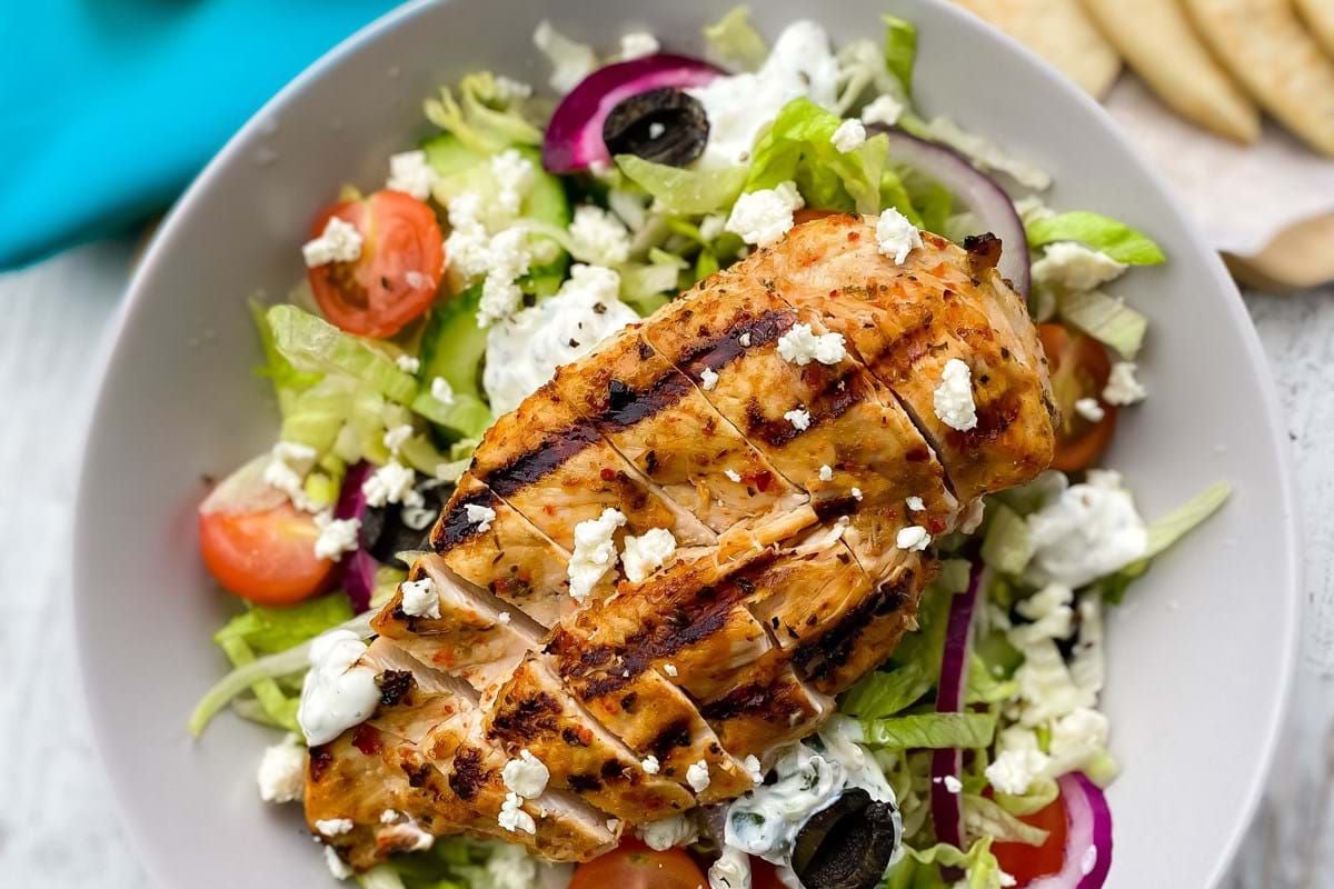 Grilled chicken breast on a salad with lettuce, tomatoes, olives, onions, and feta cheese.