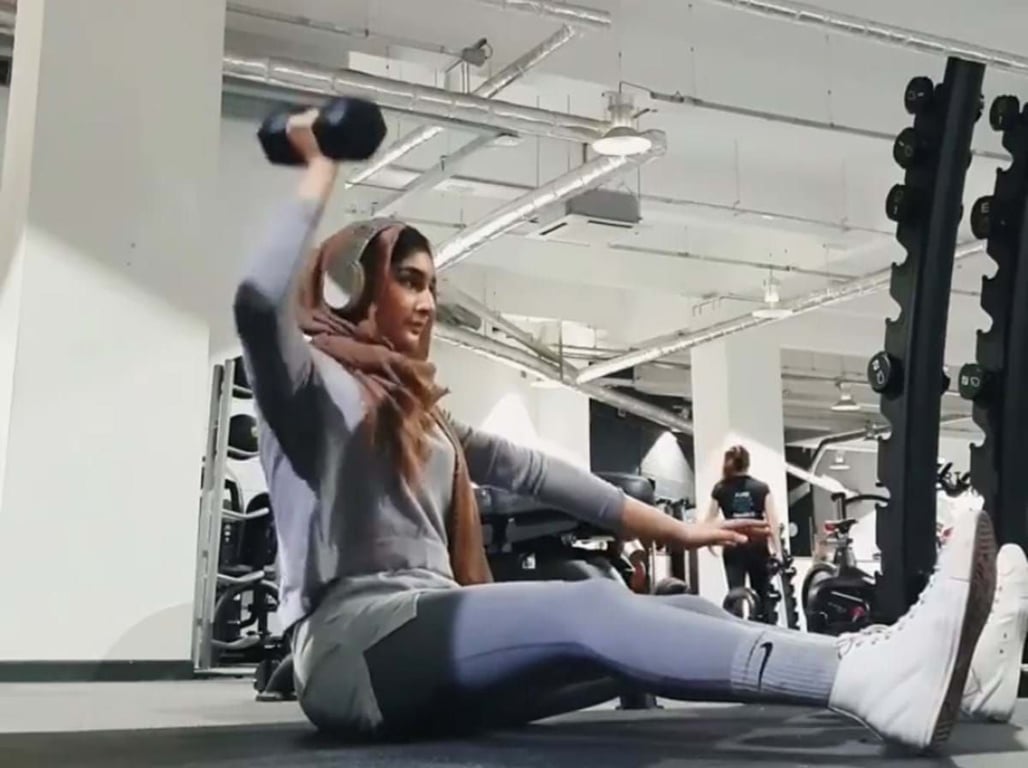 Woman in hijab lifting dumbbell while seated on gym floor with legs extended