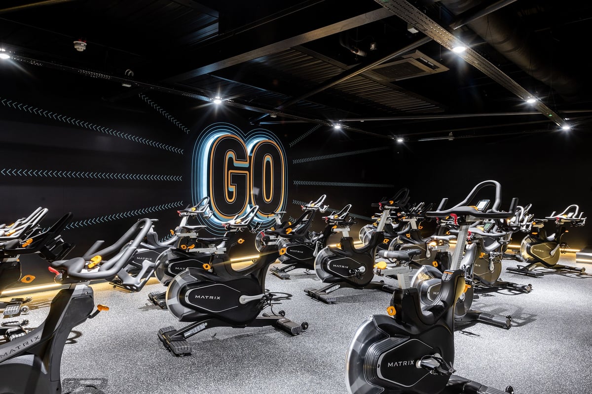PureGym Cheshunt Brookfield Shopping Park: Rows of stationary bikes in a dimly lit studio with neon "GO" sign.