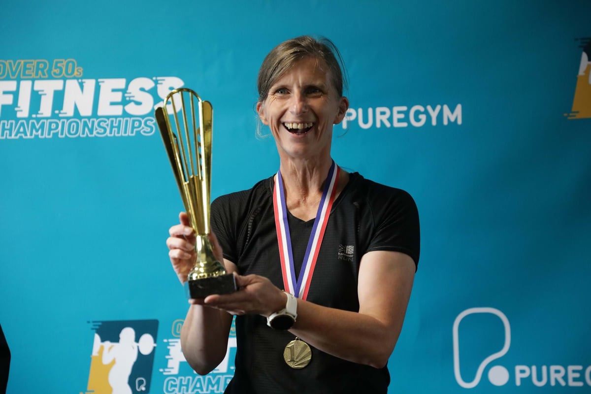 Woman in black shirt holding gold trophy and wearing gold medal at fitness event backdrop