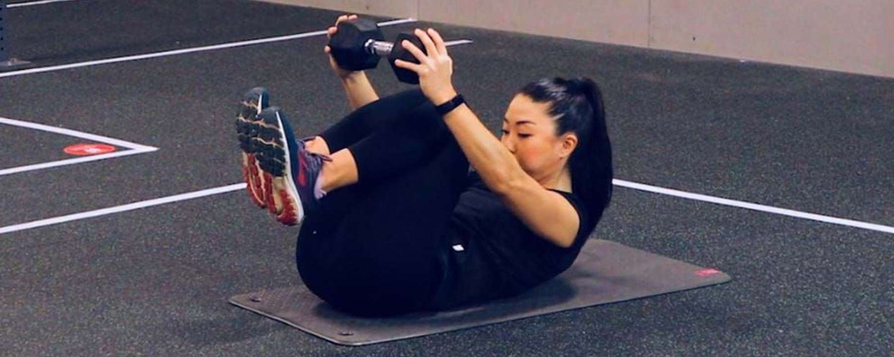 Woman in black workout clothes holding dumbbells while doing abdominal exercise on mat