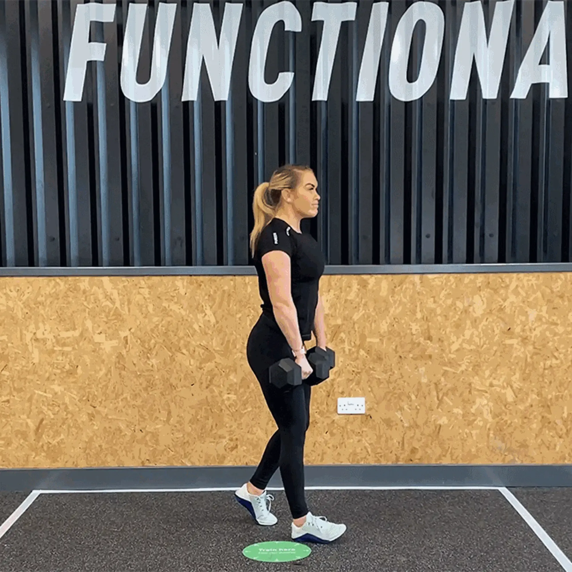 Woman in black workout clothes holding dumbbells standing on one leg in gym