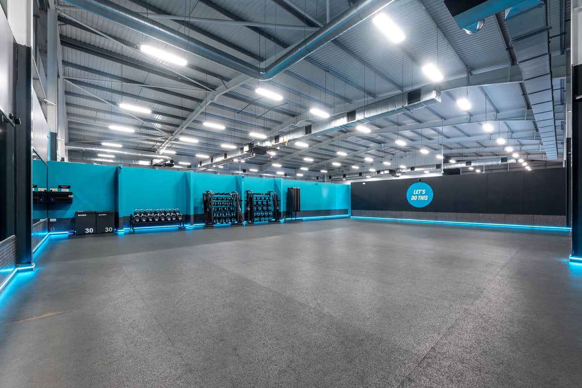 PureGym Widnes: Teal panelled walls, black rubberized flooring, weightlifting racks and equipment.