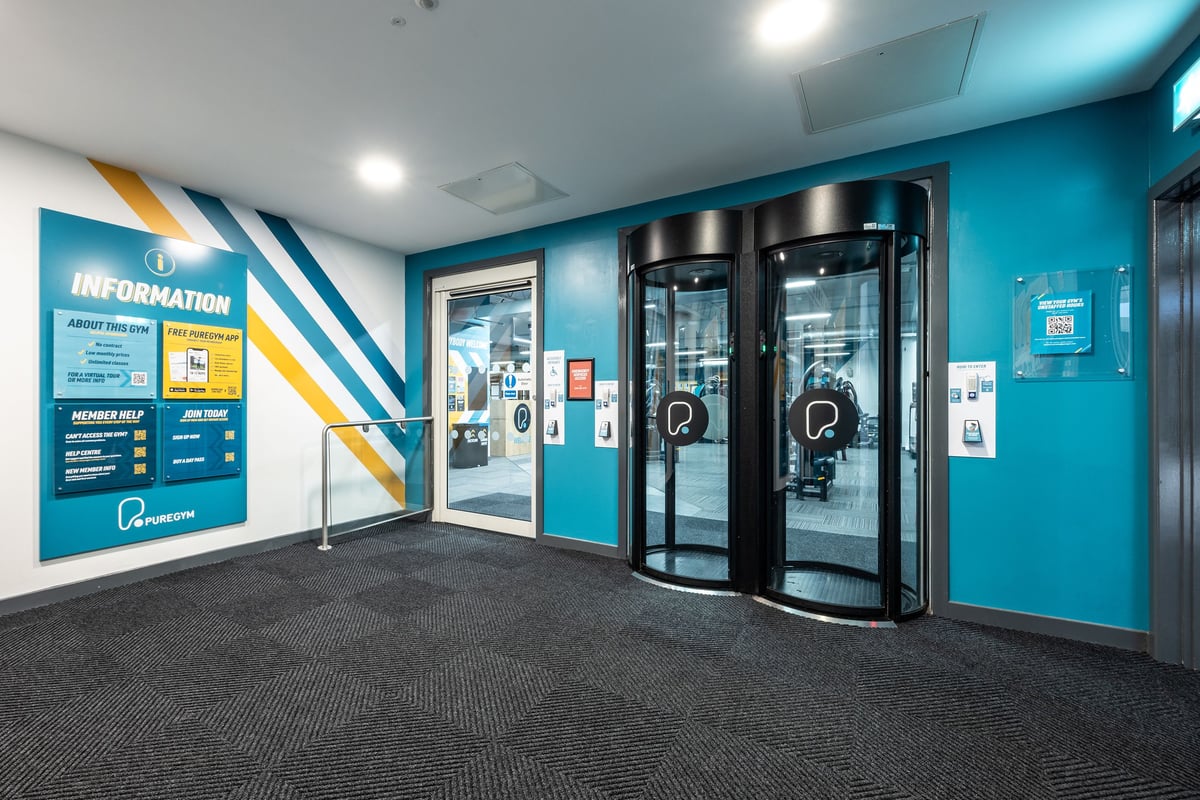 PureGym Motherwell: Information area, revolving glass doors, corporate colors of teal and yellow on walls.