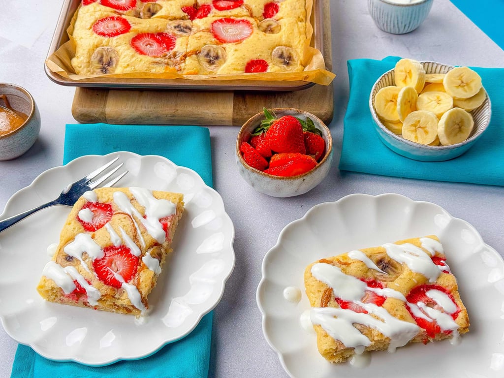 Sliced strawberry and banana cake with white icing on two plates and bowls of fruit