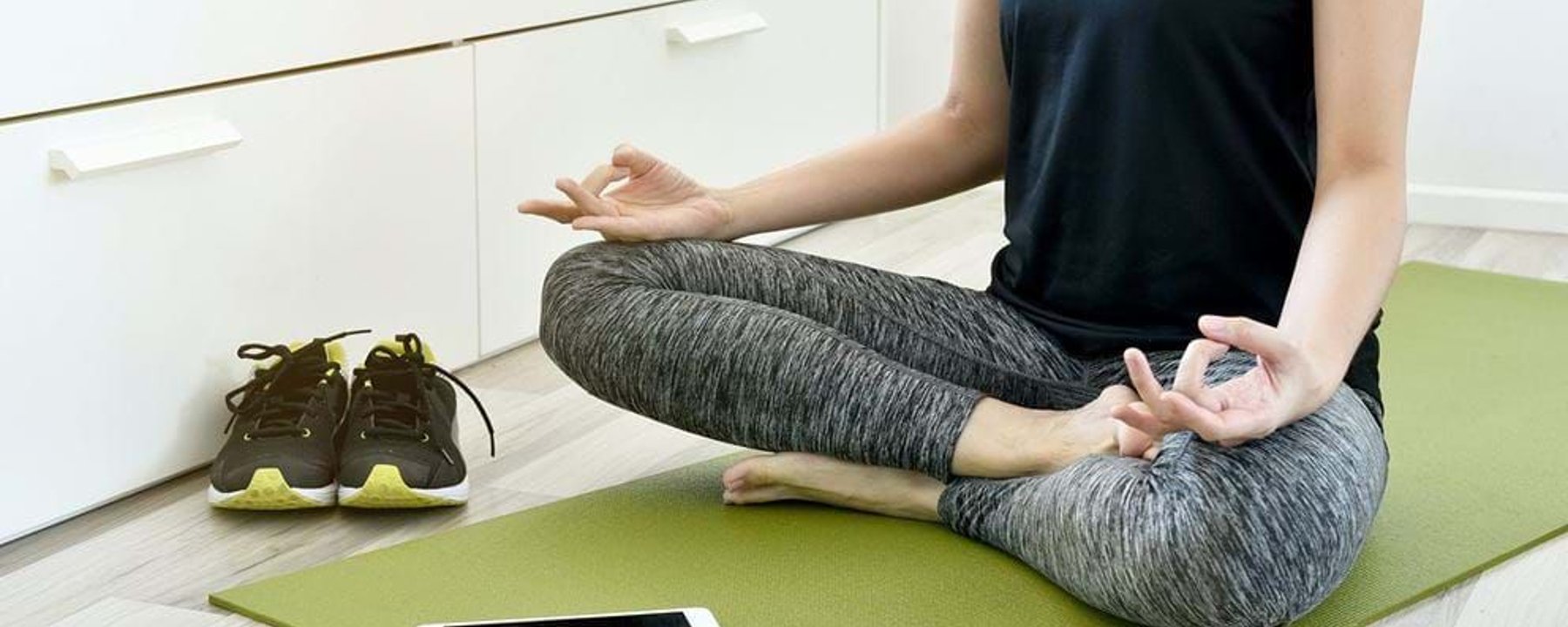 Person sitting cross-legged on a green yoga mat meditating with shoes and tablet nearby