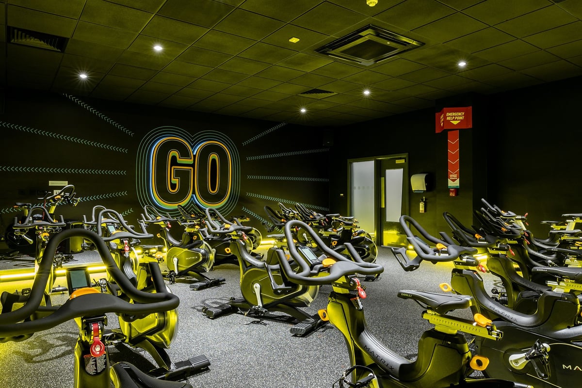 PureGym Cambridge Leisure Park: Rows of stationary exercise bikes emblazoned with neon 'GO' signage and lighting.