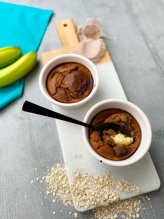 Two baked oat puddings in white ramekins on a marble board with bananas and oats nearby