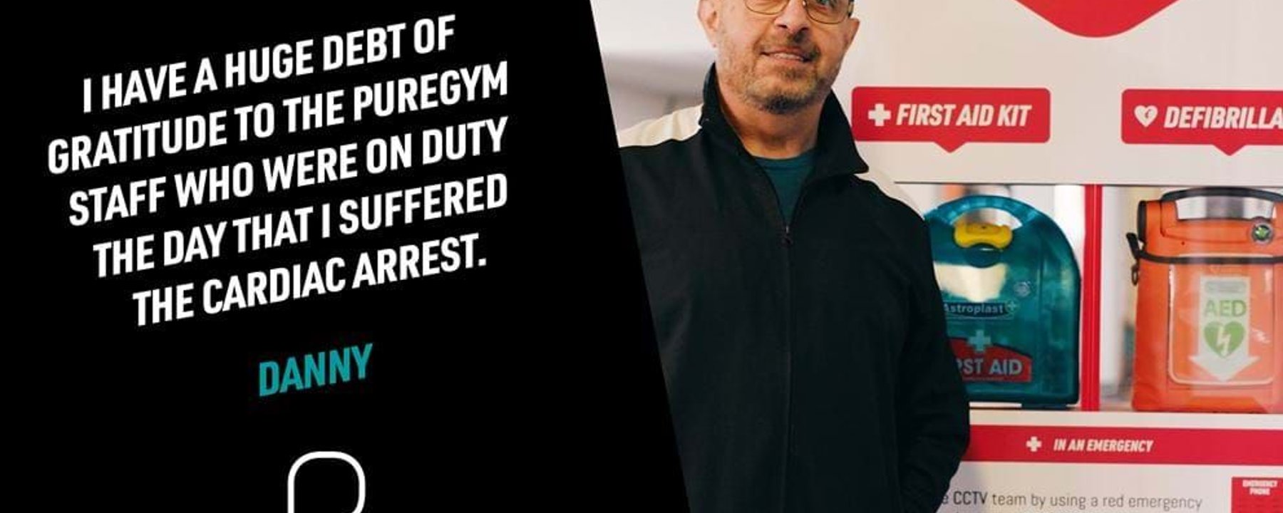 Man in glasses and black jacket stands beside a wall with first aid and defibrillator sign