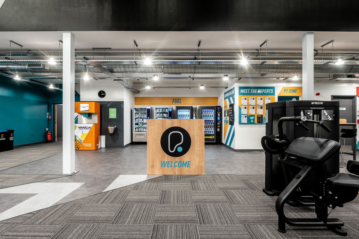 PureGym Ashford Warren Retail Park: Cardio machines, self-service kiosks, lockers, and suspended lighting in a gym facility.