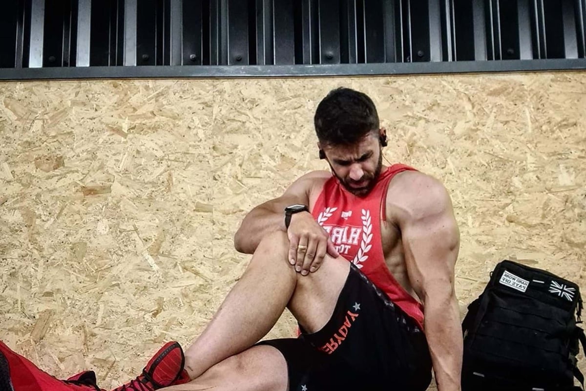 Man in red vest and black shorts sitting on gym floor stretching leg muscles