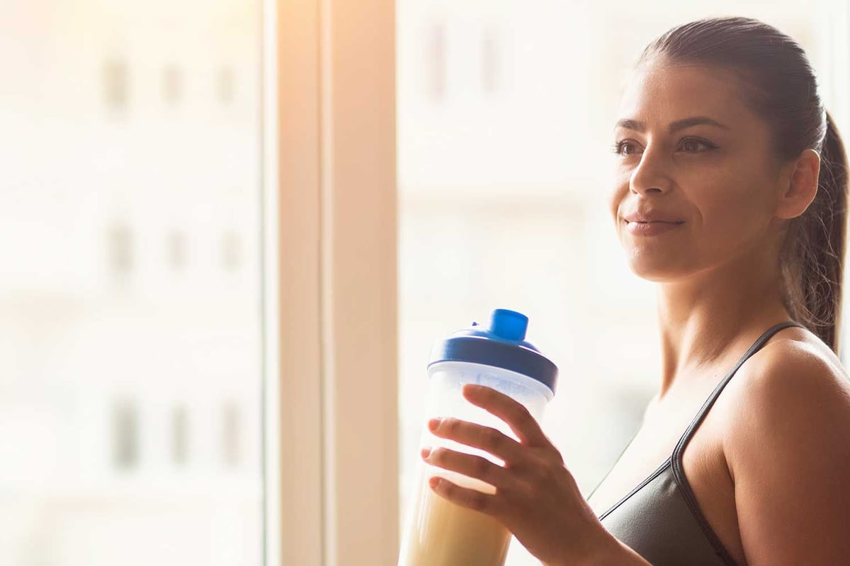 Frau in Sportkleidung mit Wasserflasche blickt aus Hochhaus-Fenster.