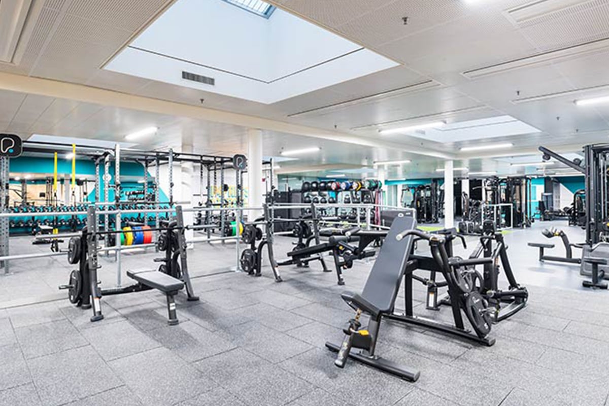 PureGym Fribourg: Salle de sport avec appareils de résistance, haltères, bancs, miroirs et sol caoutchouté sous lucarnes.