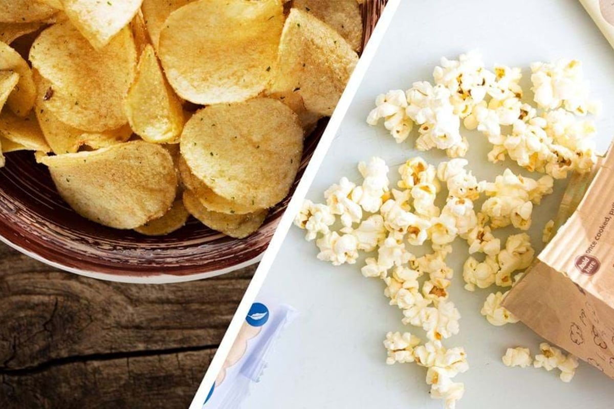 Bowl of potato chips on wooden surface and spilled popcorn from paper bag on white surface
