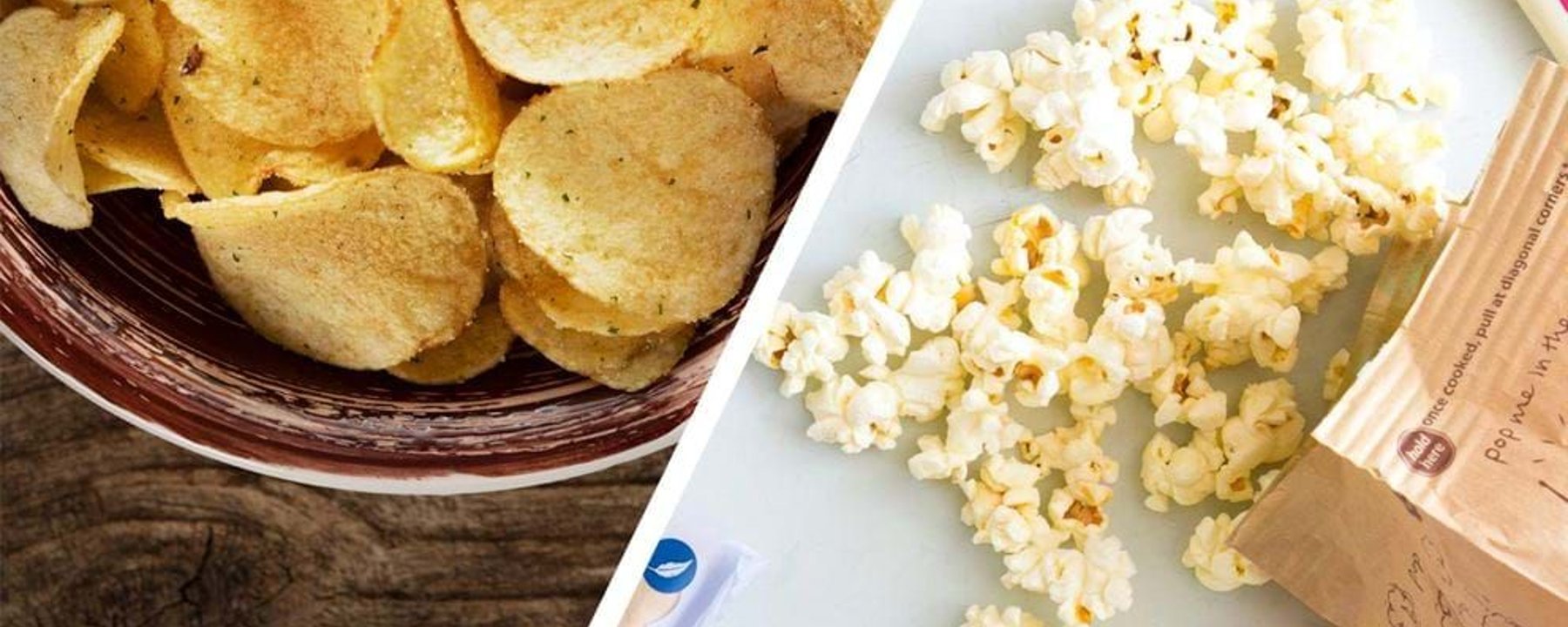 Bowl of potato chips on wooden surface and spilled popcorn from paper bag on white surface