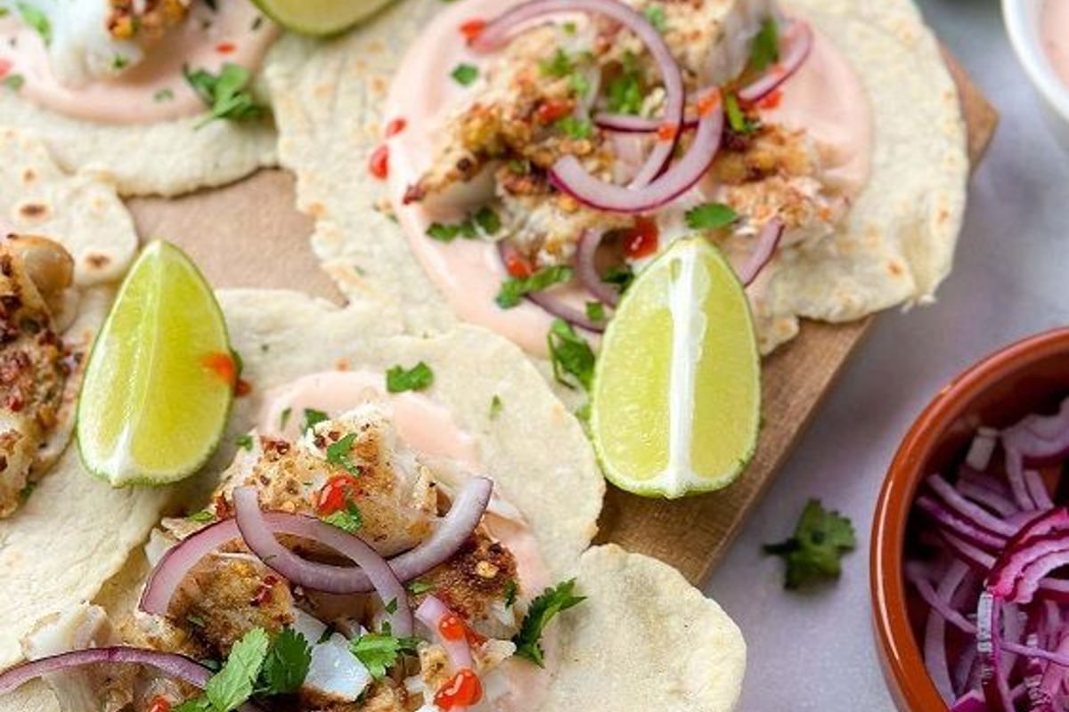 Tortillas topped with grilled fish, red onion, sauce, herbs, and lime wedges on a board