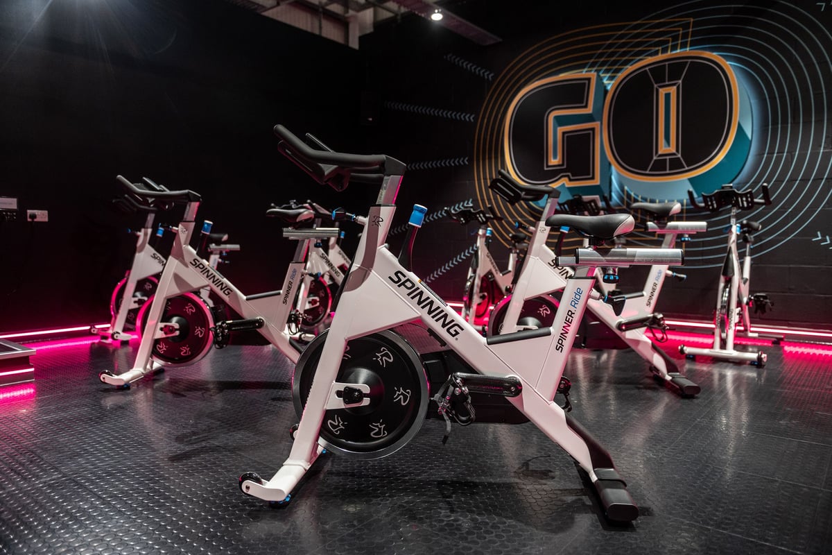 PureGym Swindon Stratton: Rows of stationary bikes, illuminated by neon lights and logos in a dark studio setting.