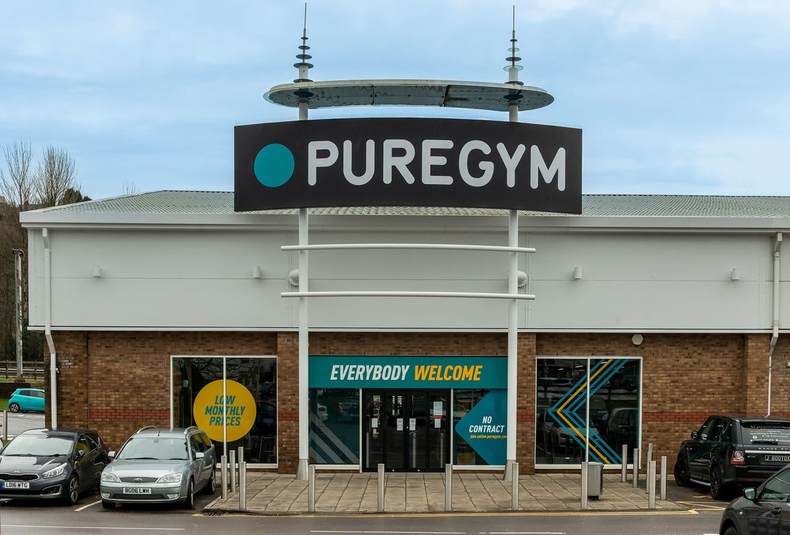 PureGym Llantrisant: PureGym storefront with branded signage, parked cars, and 'Everybody Welcome' window displays.