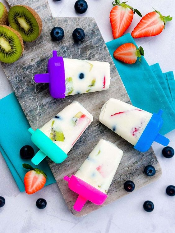 Frozen yoghurt popsicles with kiwi, strawberry, and blueberry on a marble board.