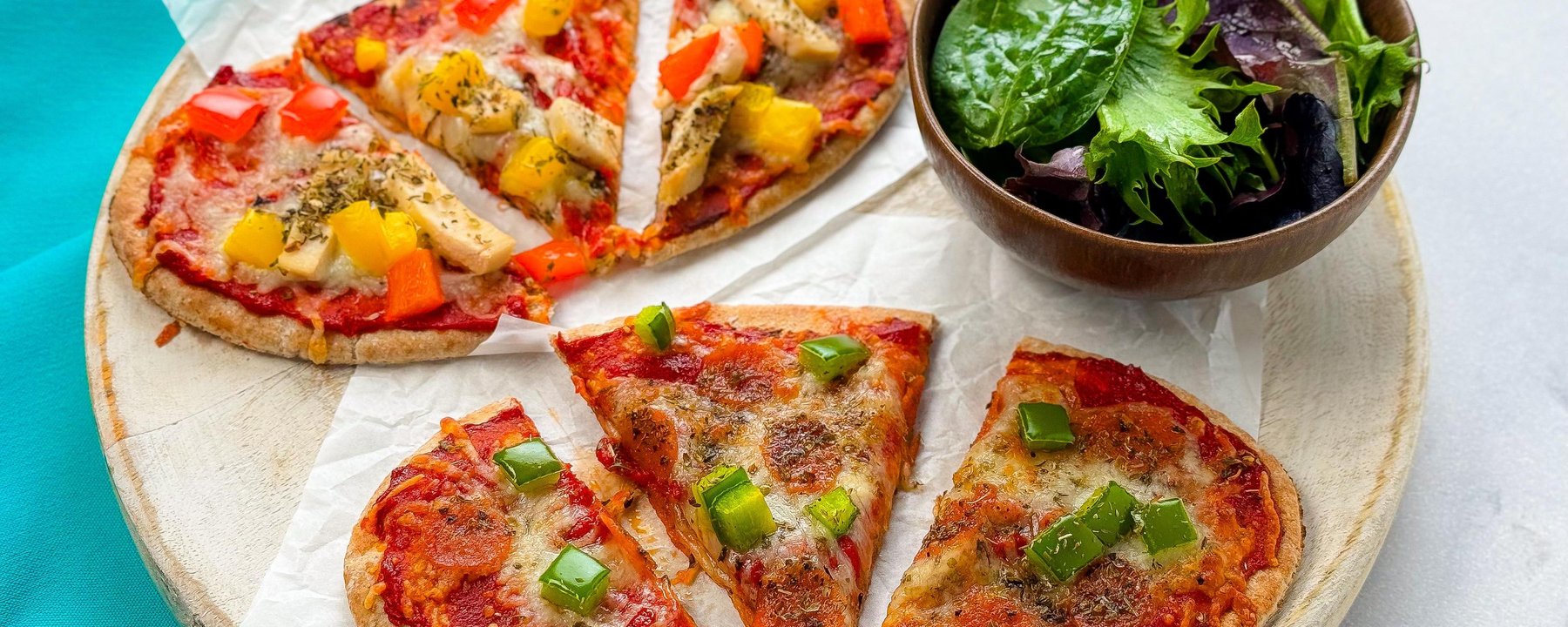 Pizza slices on wooden board with colourful toppings served with bowl of salad leaves