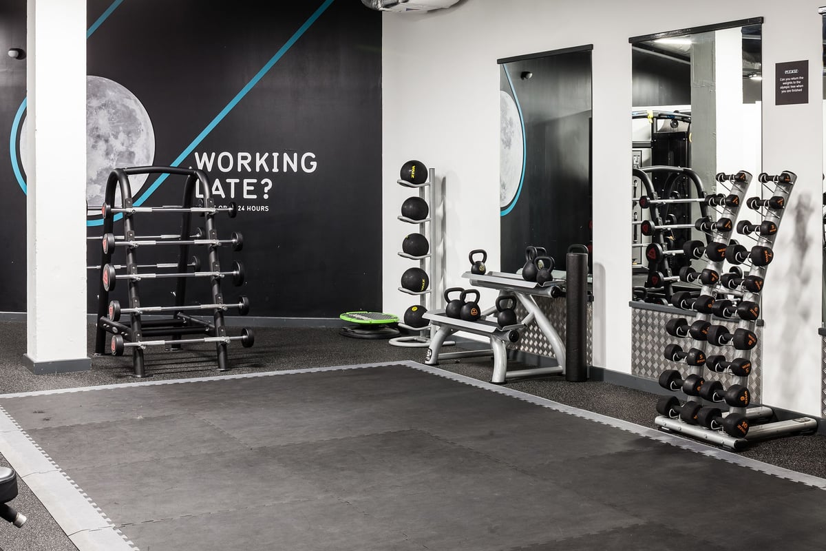 PureGym Manchester Stretford: Weightlifting racks, free weights, benches, and mirrored walls in a dimly lit gym space.