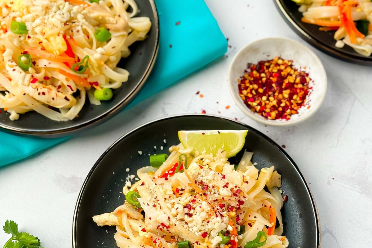 Black plates with noodle salad garnished with lime wedges, peanuts, and spring onions