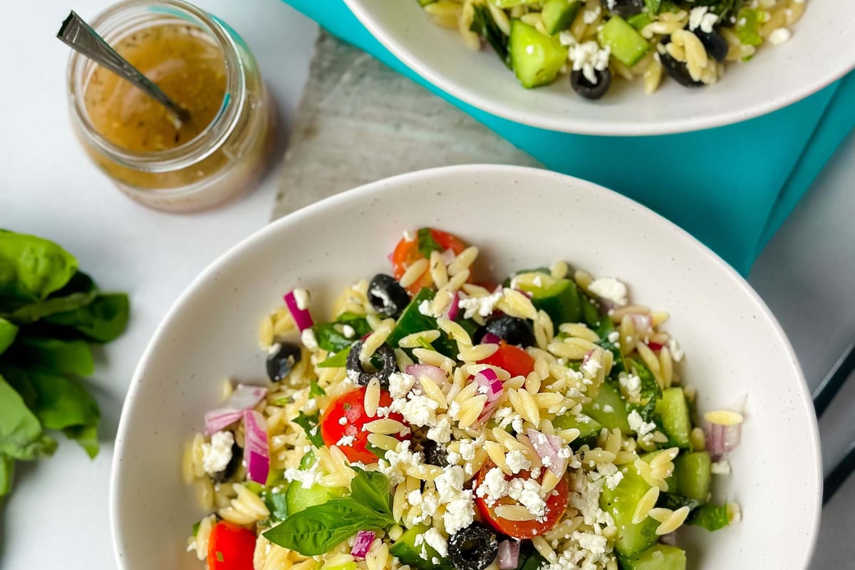 Two bowls of orzo salad with cherry tomatoes, cucumber, olives, feta, and herbs