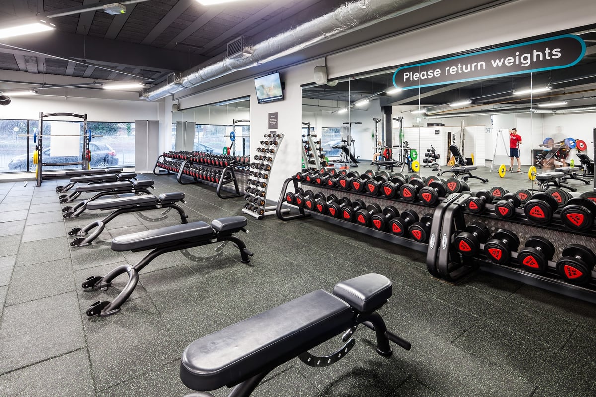 PureGym Manchester Stretford: Weight racks, free weights, benches, and rubberised flooring in a gym weight room.