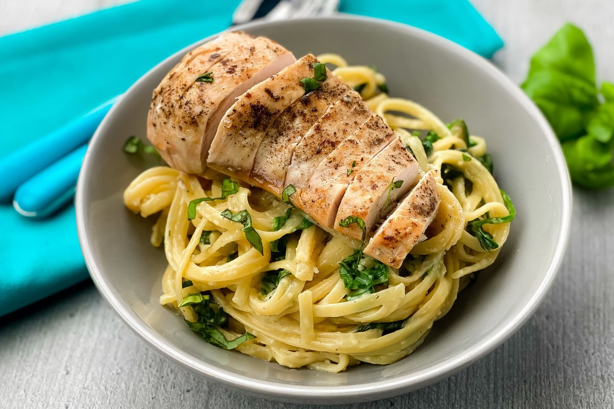 Sliced grilled chicken breast on pasta with herbs in a white bowl