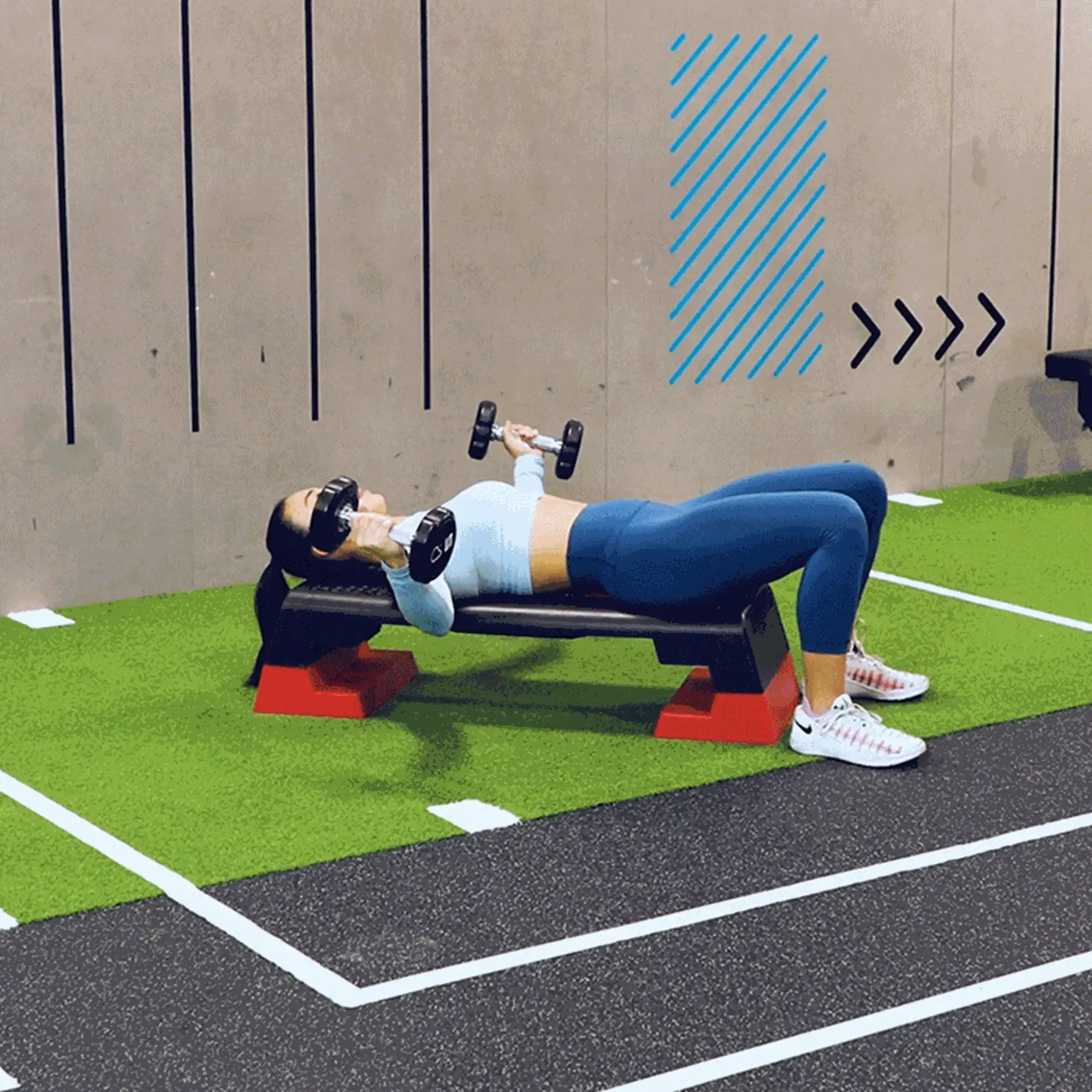 Woman in blue leggings lying on bench lifting dumbbells in gym with green turf.