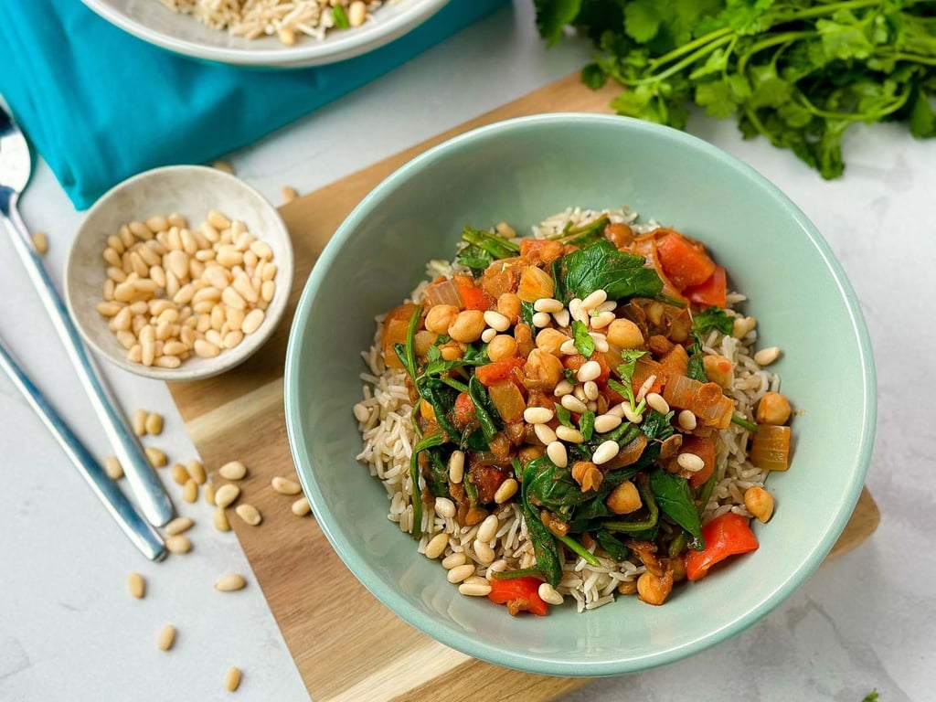 Bowl of rice topped with chickpeas, spinach, red peppers, and pine nuts on a wooden board