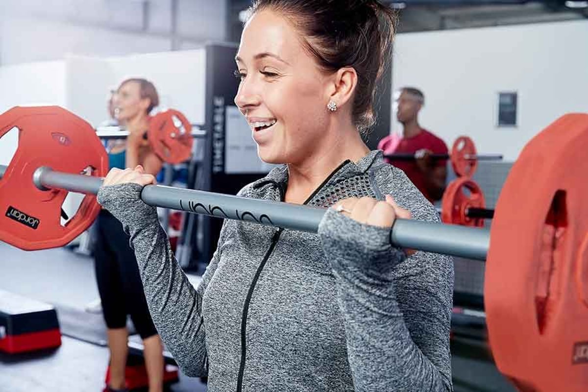Frau hebt Langhantel mit roten Gewichten, lächelt beim Training mit anderen im Hintergrund