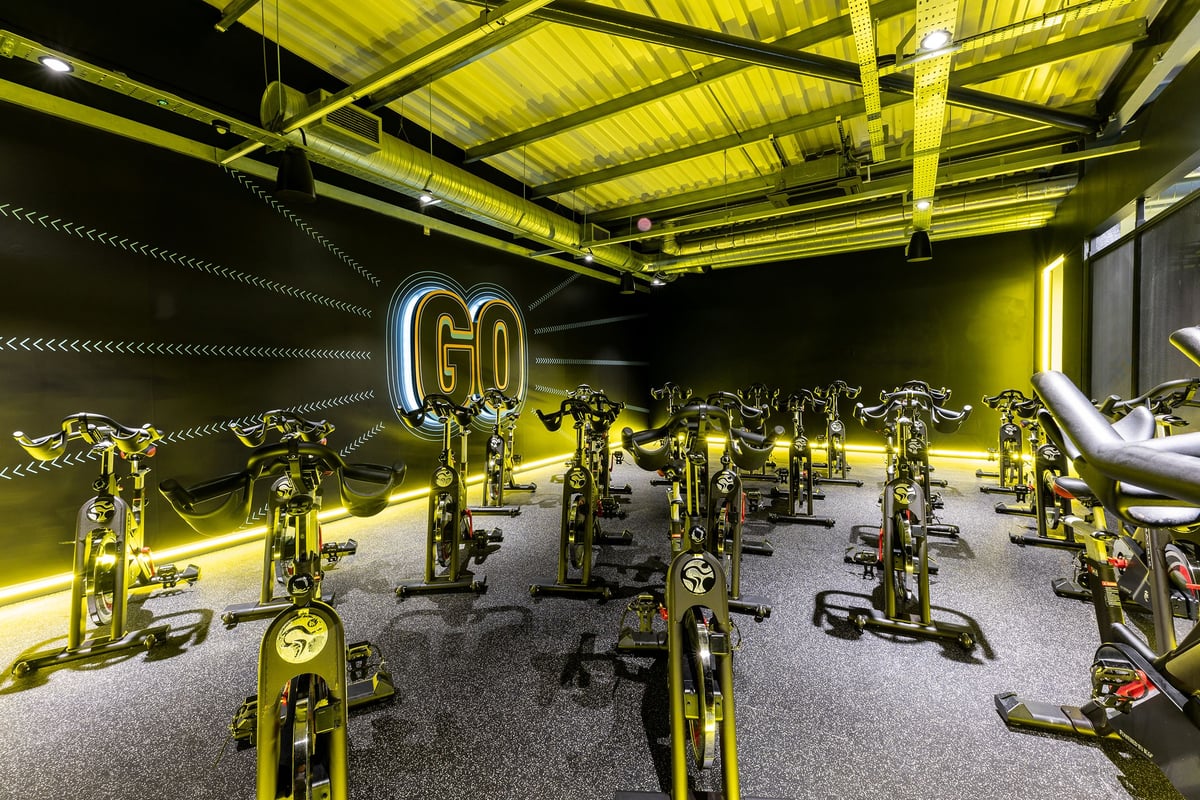 PureGym Reading Basingstoke Road: Rows of stationary cycles, illuminated neon signage, industrial ceiling with exposed beams.