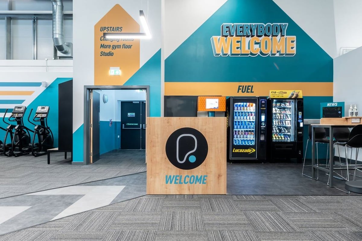 PureGym Kettering: Vending machines, snack racks, seating areas with "Everybody Welcome" signage indicating a gym lobby area.
