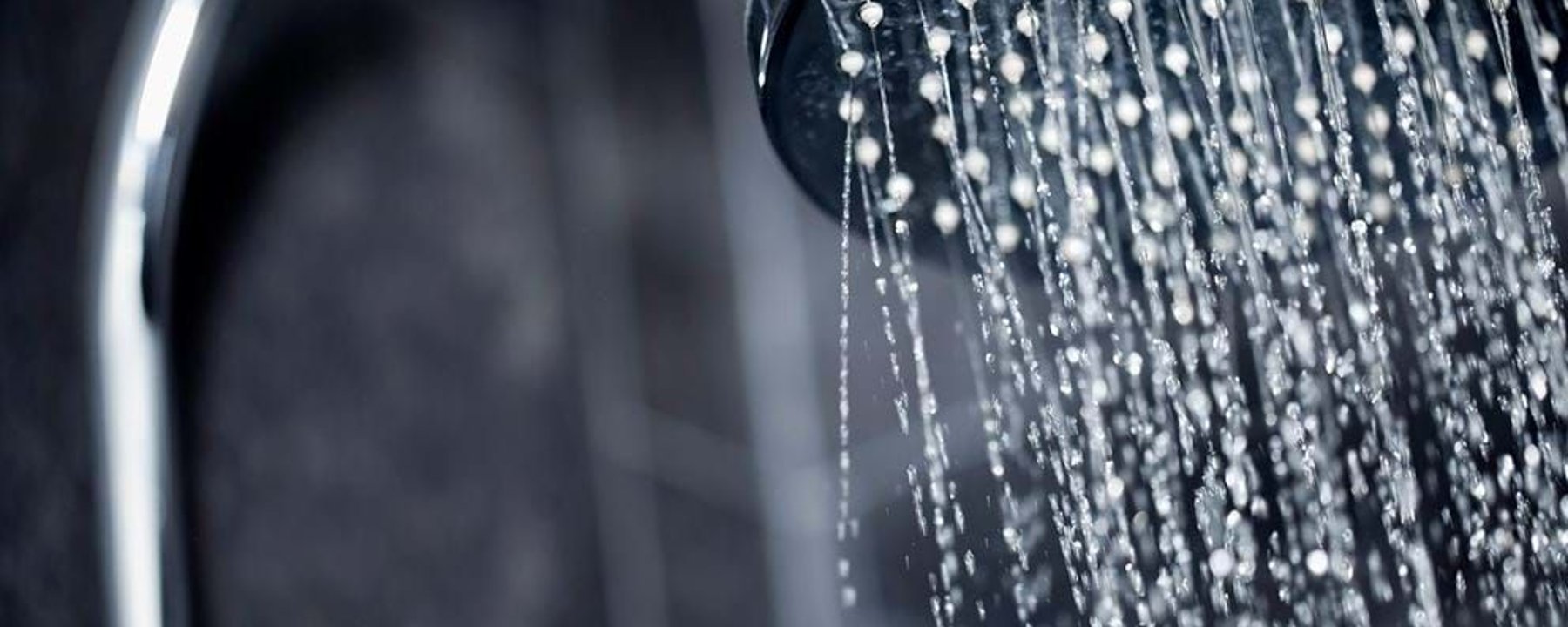 Water flowing from a round metal showerhead against a dark tiled wall