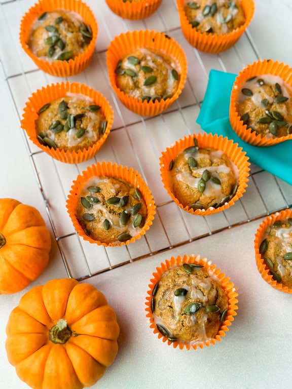 Pumpkin muffins with glaze and seeds in orange liners on a cooling rack with pumpkins