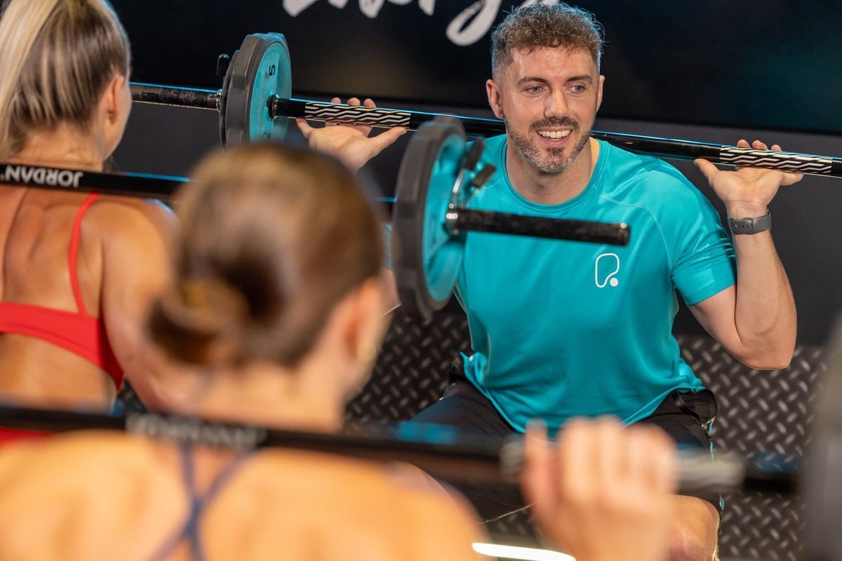 Male instructor in turquoise shirt leading group fitness class with weighted bars