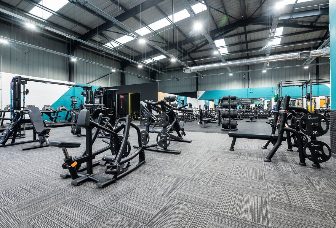 PureGym Dunfermline: Rows of seated weightlifting machines, free weights, and rubberized flooring in a spacious gym area.
