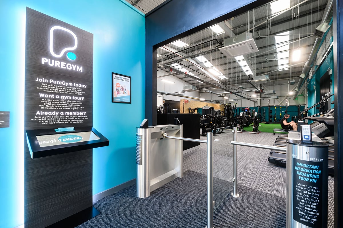 PureGym Buckingham: Reception area with self-service kiosks, turnstiles, and weight training equipment visible beyond.