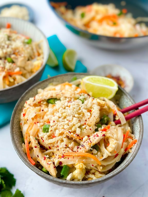 Bowl of pad thai noodles with crushed peanuts, lime wedge, and red chopsticks.