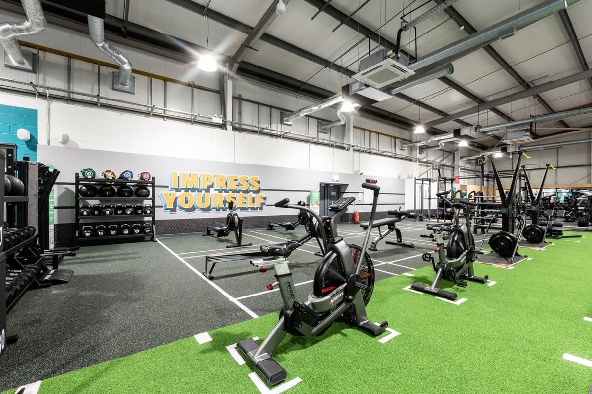 PureGym Swindon Stratton: Resistance machines, exercise bikes, free weights, and green turf in a spacious gym interior.