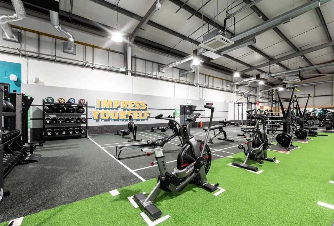 PureGym Swindon Stratton: Resistance machines, exercise bikes, free weights, and green turf in a spacious gym interior.
