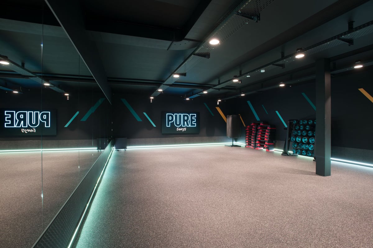 PureGym Adliswil: Studio de gym sombre avec la marque Neon Pure, des arbres à haltères, un sol caoutchouté et des murs en miroir.