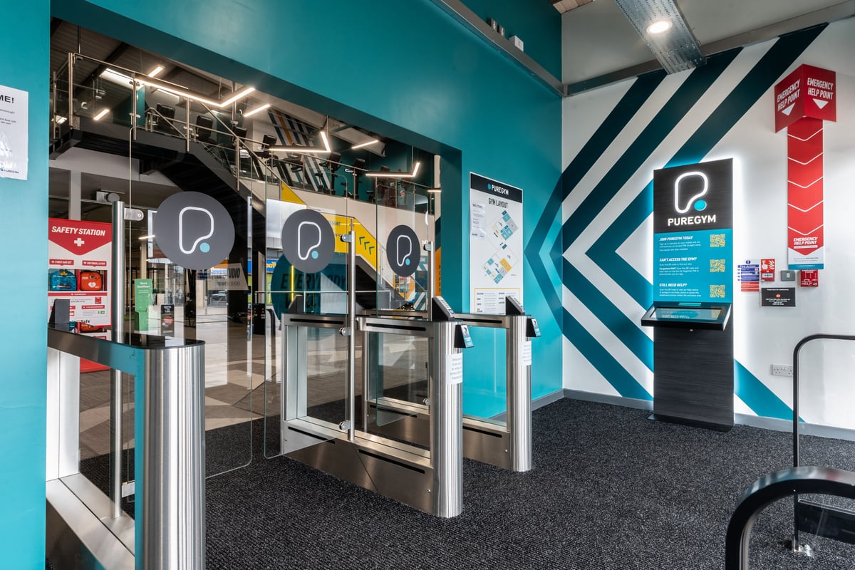PureGym Peterborough Brotherhood Retail Park: Turnstiles, electronic kiosks, floor signage indicating directions.