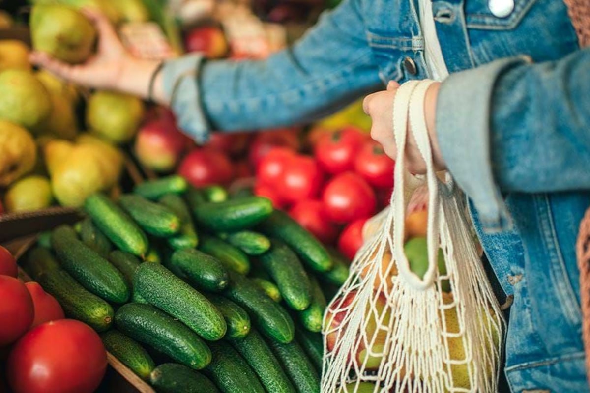 Person in denim jacket picking fruit at market stall with cucumbers and tomatoes