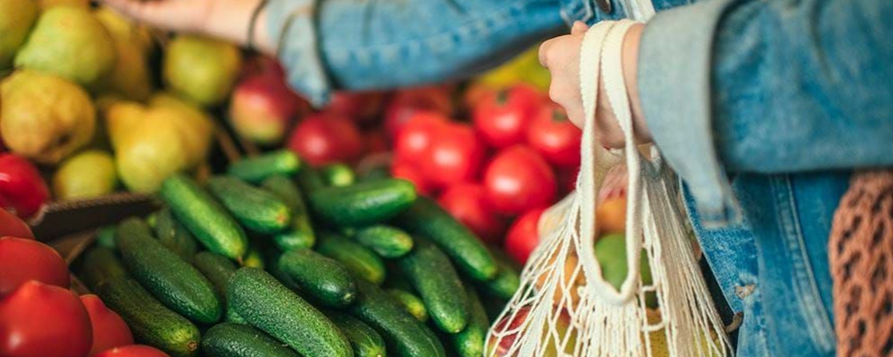 Person in denim jacket picking fruit at market stall with cucumbers and tomatoes