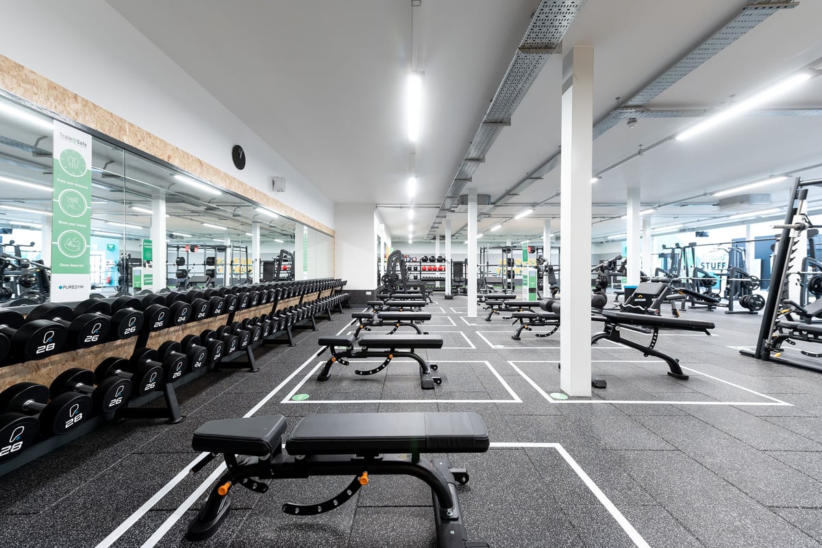 PureGym Colchester Retail Park: Gym interior with weight benches, dumbbell racks, mirrored walls, and rubberized flooring.