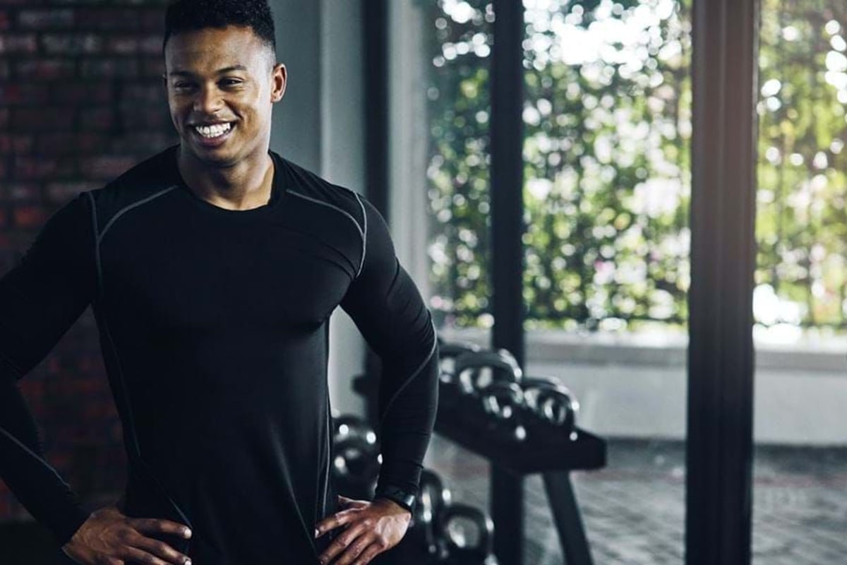 Smiling man in black sportswear standing with hands on hips in gym near kettlebells.