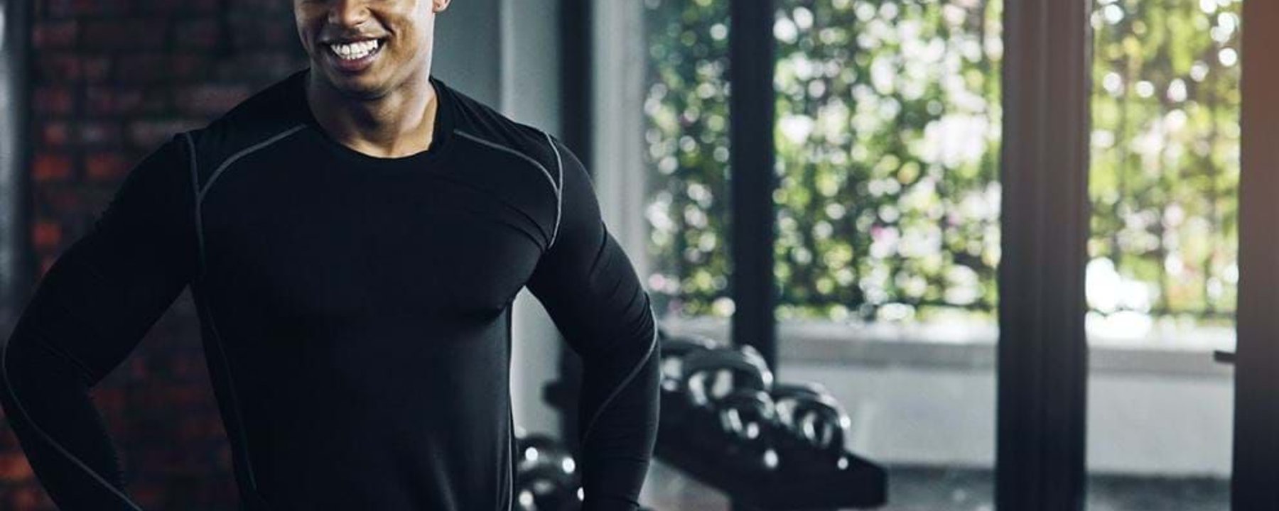 Smiling man in black sportswear standing with hands on hips in gym near kettlebells.