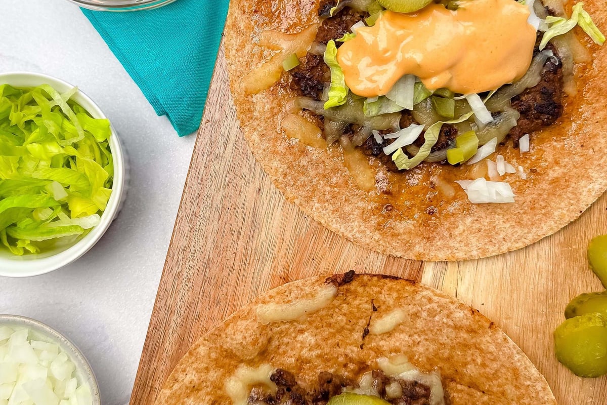 Two open wraps with beef, cheese, pickles, lettuce, onions, and sauce on a wooden board.